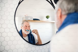 A senior man brushing teeth in bathroom indoors at home, looking in mirror. Copy space.