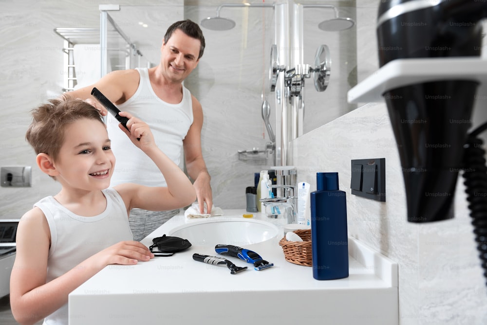 Waist up photo of smiling father and happy little son standing together while boy combing hair and father looking on him