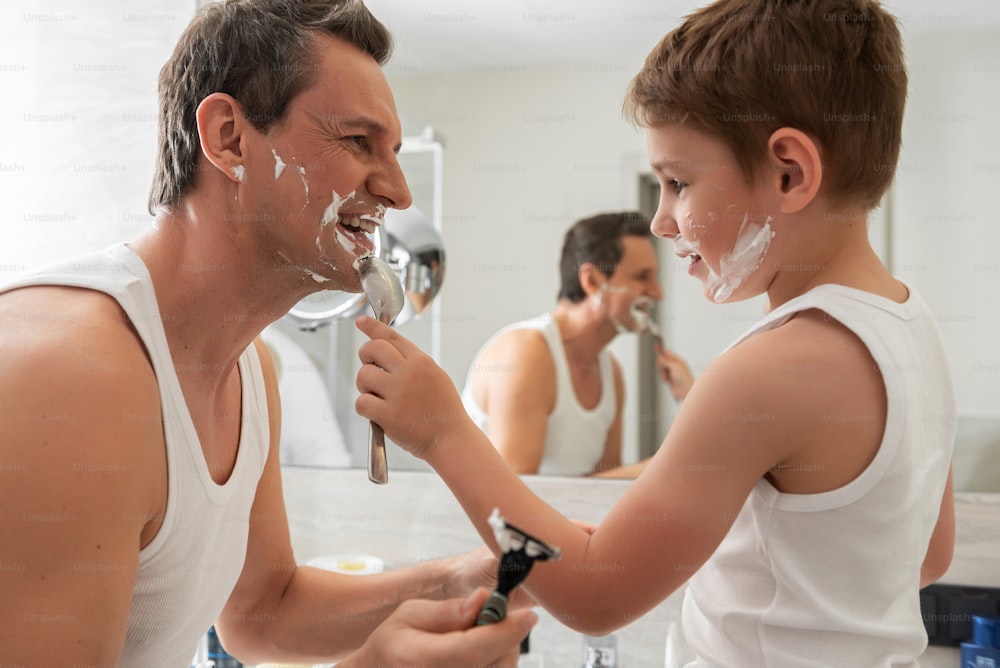Waist up portrait of smiling father and happy little son wearing white T-shirts and holding razors near mirror