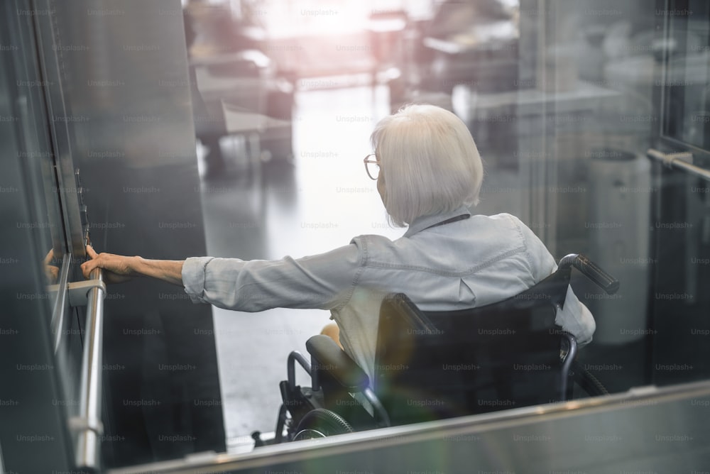 Foto da vista de trás da velha senhora com bolsa branca e sentada em cadeira de rodas enquanto pressiona o botão do elevador