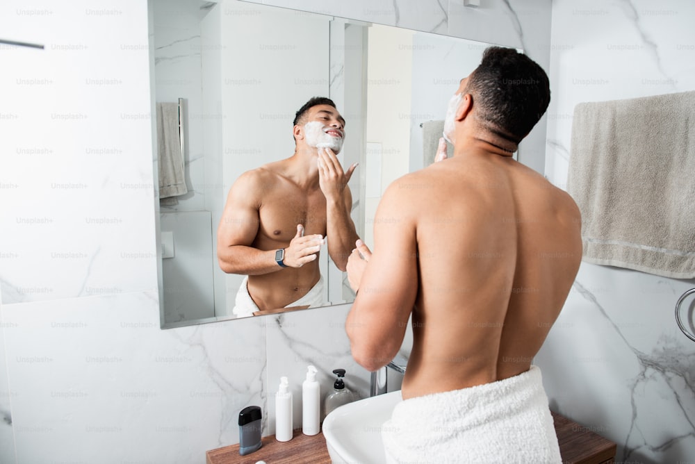 Morning hygiene concept. Waist up back side portrait of young naked muscular male spreading shaving foam on face while staying before mirror in bathroom