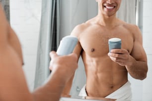 Morning hygiene concept. Waist up cropped head reflection of young cheerful naked muscular male holding antiperspirant while staying before mirror in bathroom
