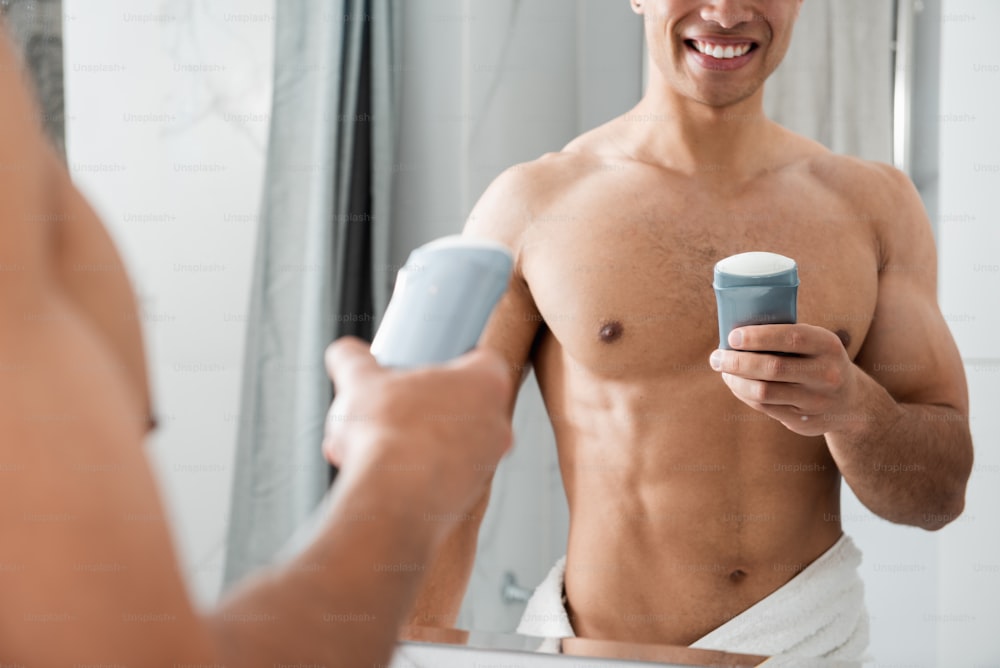 Concept d’hygiène matinale. Taille vers le haut tête recadrée reflet d’un jeune homme musclé nu joyeux tenant un antisudorifique tout en restant devant le miroir dans la salle de bain