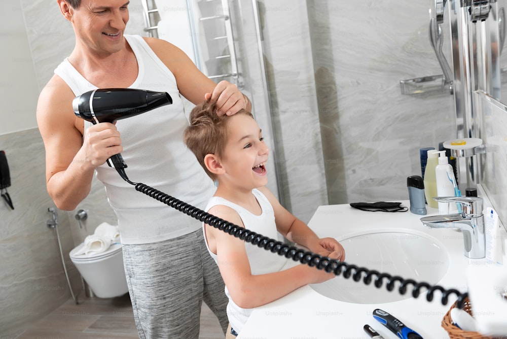 Taillenporträt des lächelnden Vaters, der dem Sohn die Haare schneidet, während er den Haartrockner benutzt und zu Hause in der Nähe des Spiegels steht