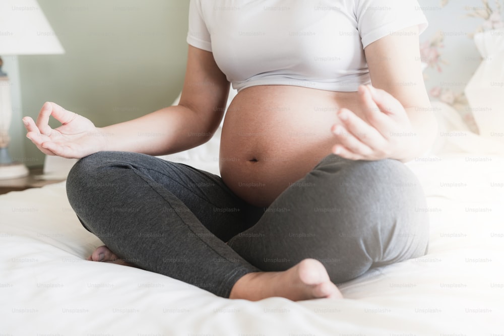 임신한 여자는 아이를 돌보면서 집 침실의 침대에서 요가 운동을 하고 있다. 임신한 뱃속에 아기를 안고 있는 행복한 젊은 임산부. 출산 산전 관리 및 임신.