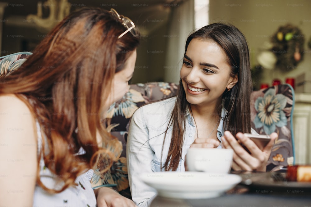 Beautiful young caucasian woman with dark long hair looking at her female friend smiling while holding a smartphone while sitting in restaurant.