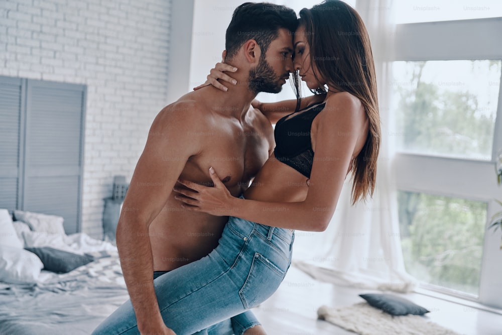 Hombre joven guapo sin camisa que lleva a una mujer atractiva semivestida mientras está de pie en el dormitorio