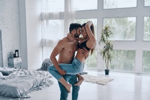 Handsome young shirtless man carrying semi-dressed attractive woman while standing in the bedroom