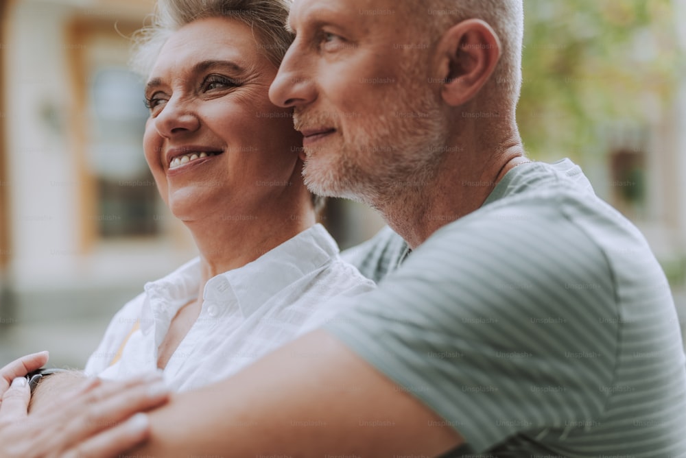 Waist up portrait of positive mature male and female in casual wear hugging outdoors while looking away