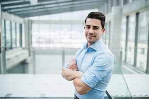 A portrait of young businessman standing indoors in an office, arms crossed. Copy space.