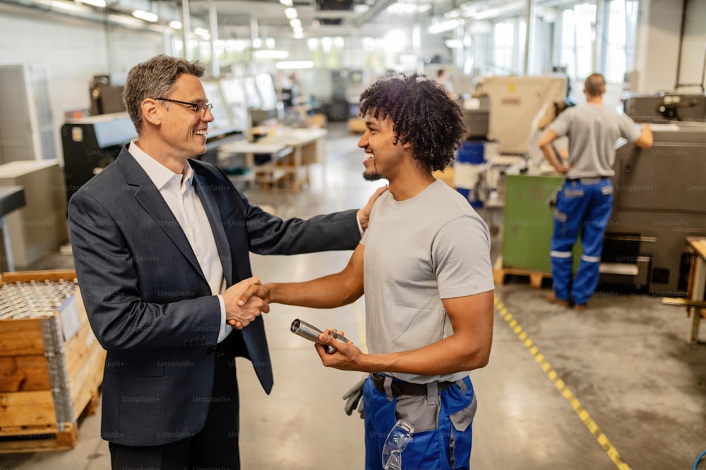 Happy manager greeting with African American manual worker and shaking hands with him in factory plant.