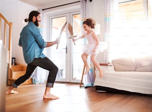 A small girl and young father with paper swords at home, fighting and playing.