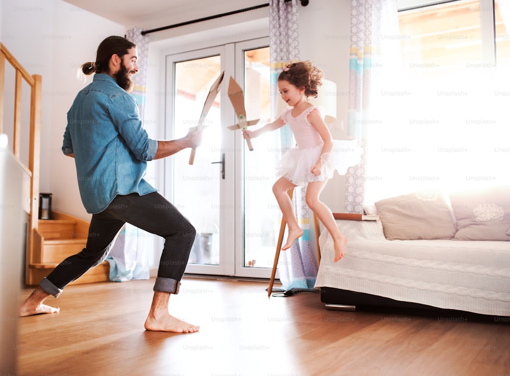 Una niña pequeña y un padre joven con espadas de papel en casa, peleando y jugando.