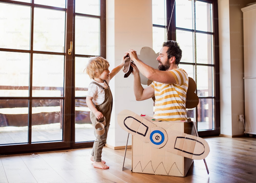 A happy toddler boy and his father with carton plane playing indoors at home, flying concept.