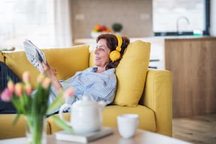 A senior woman with headphones and crosswords sitting on sofa indoors at home, resting.