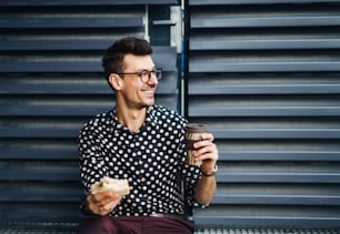 A young businessman sitting outdoors, drinking coffee and eating sandwich. Copy space.
