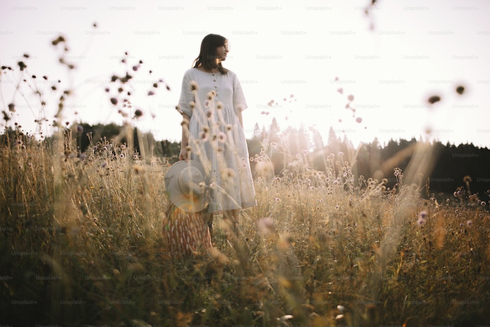 Menina elegante no vestido rústico em pé entre flores silvestres e ervas no prado ensolarado nas montanhas. Mulher Boho relaxando no campo ao pôr do sol, vida simples. Imagem atmosférica. Texto com espaço