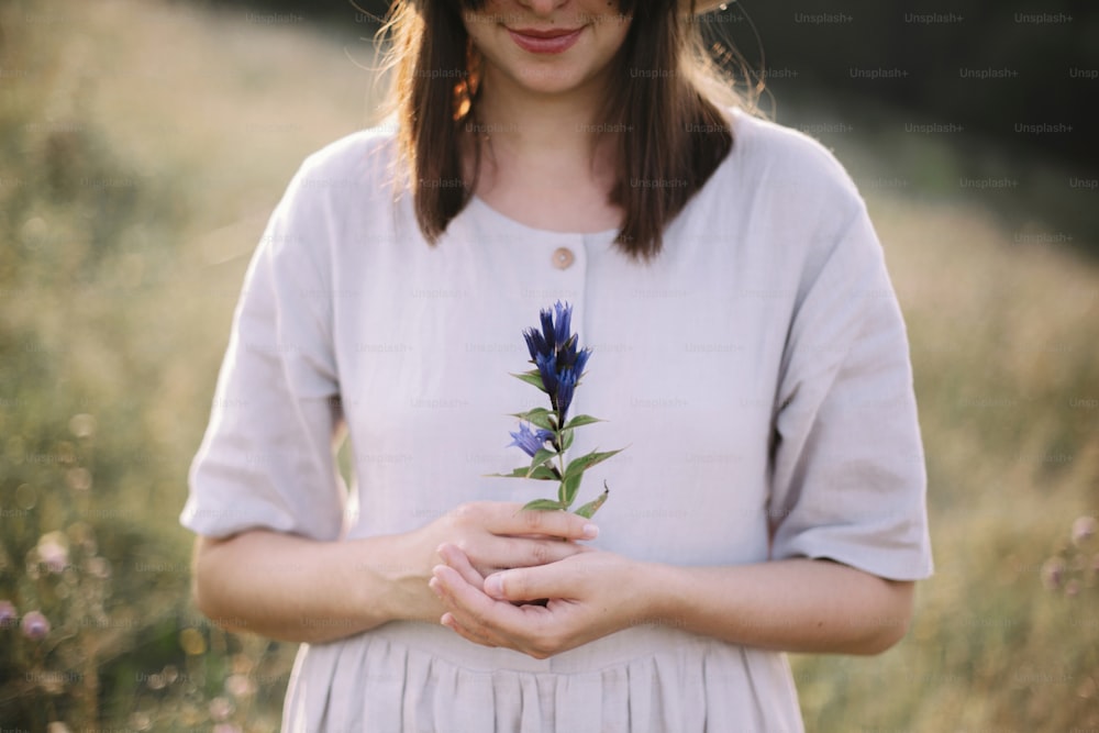 Stylish girl in rustic dress holding wildflower in hand, standing in sunny meadow in mountains. Boho woman gathering gentiana flowers in countryside, rural simple life. Atmospheric image