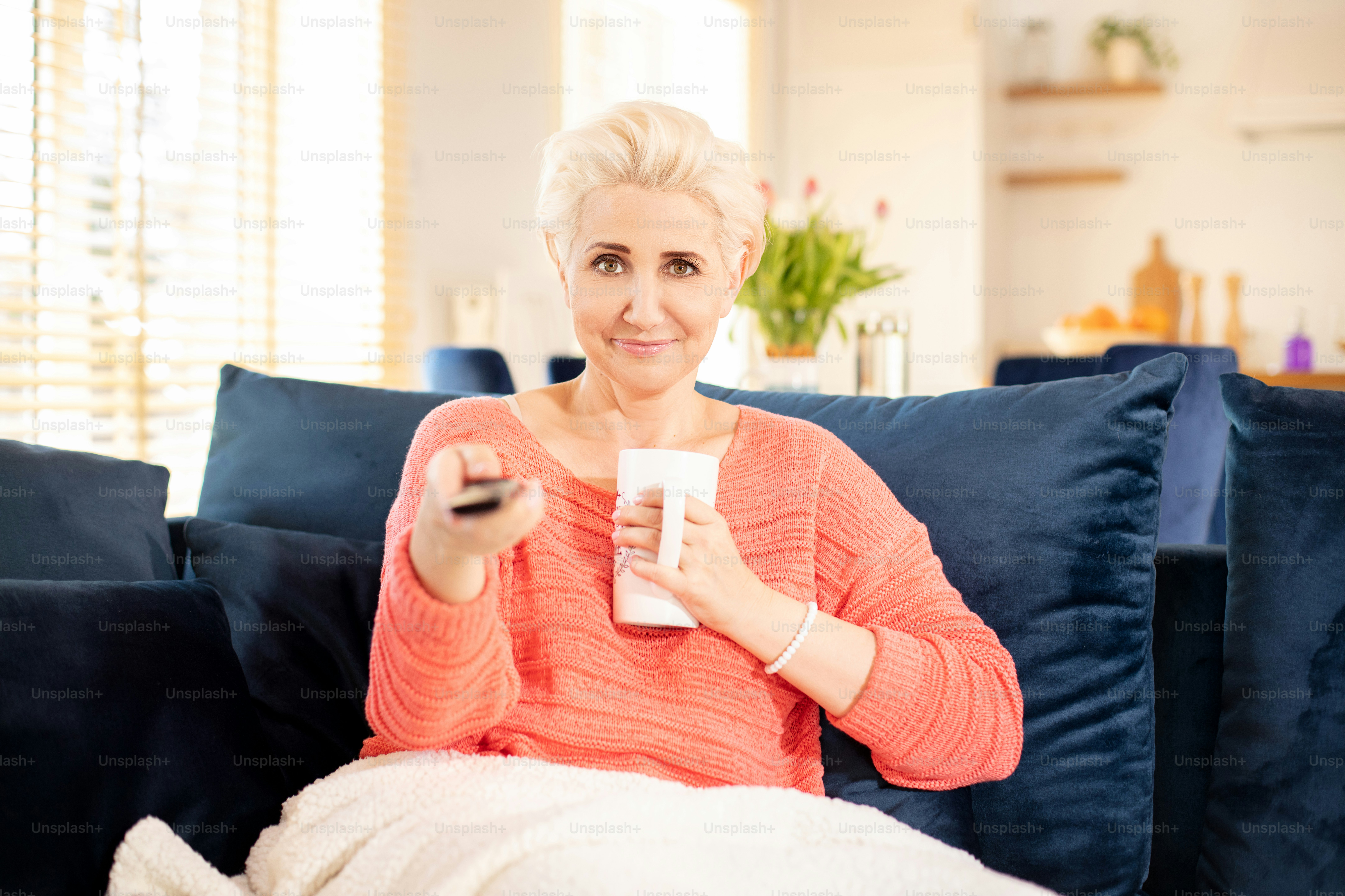 Wanita dewasa bahagia yang cantik bersantai di rumah, memegang remote control dan menonton tv.