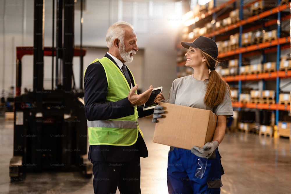 Happy female warehouse employee communicating with senior businessman while carrying cardboard box.
