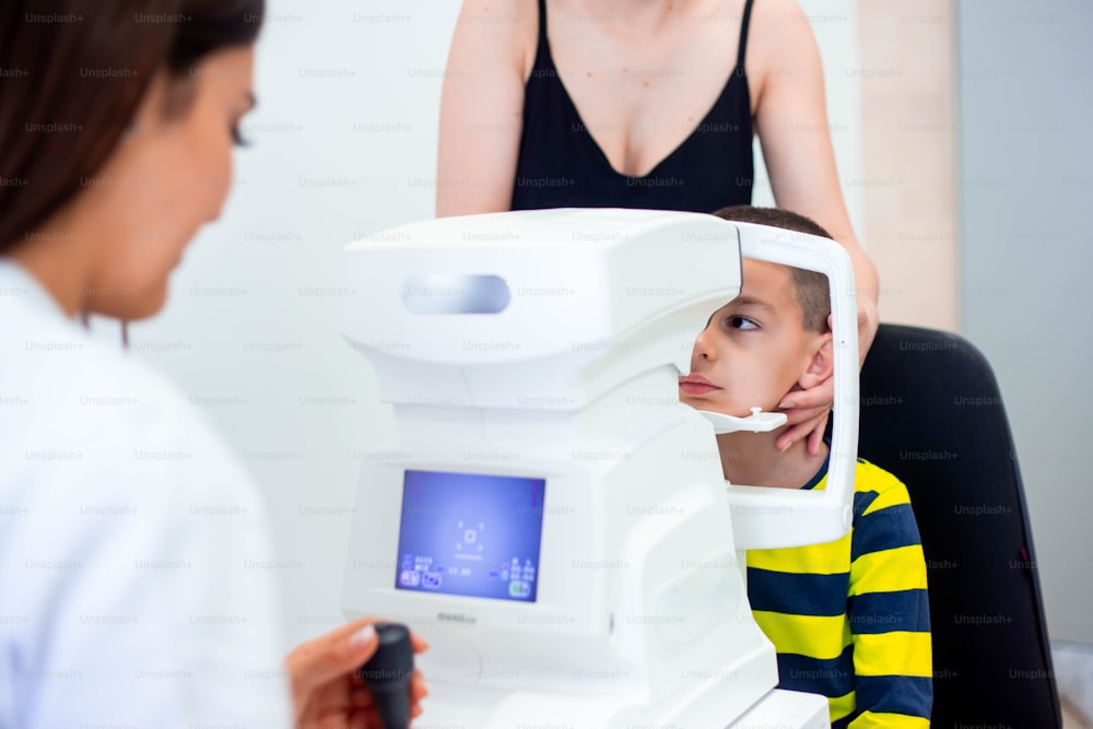 Weibliche Augenärztin, die eine Maschine zur Überprüfung des Sehvermögens in der Klinik verwendet. Kleiner Junge, der sich Geräte anschaut und Augenpupille im Optikergeschäft testet. Konzept der Augenpflege und Gesundheit