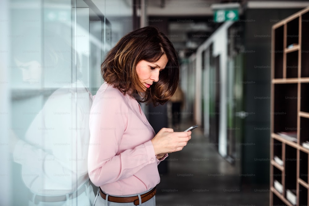 A portrait of businesswoman with smartphone standing in an office building, text messaging.