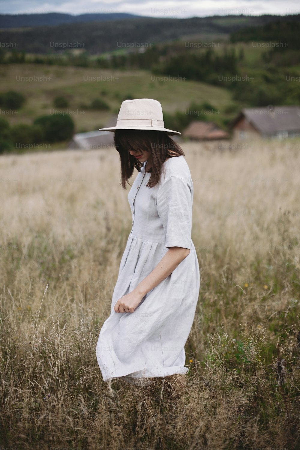 Stylish girl in linen dress and hat walking among herbs and wildflowers in field. Boho woman relaxing in countryside, simple slow life style. Space for text. Atmospheric image