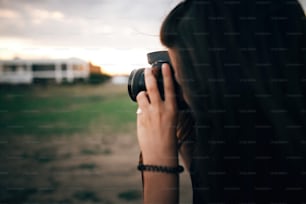 Stylish hipster girl taking photos on beach at sunset. Summer vacation. Portrait of  happy boho woman holding photo camera and making photos in evening time at  sea cliff