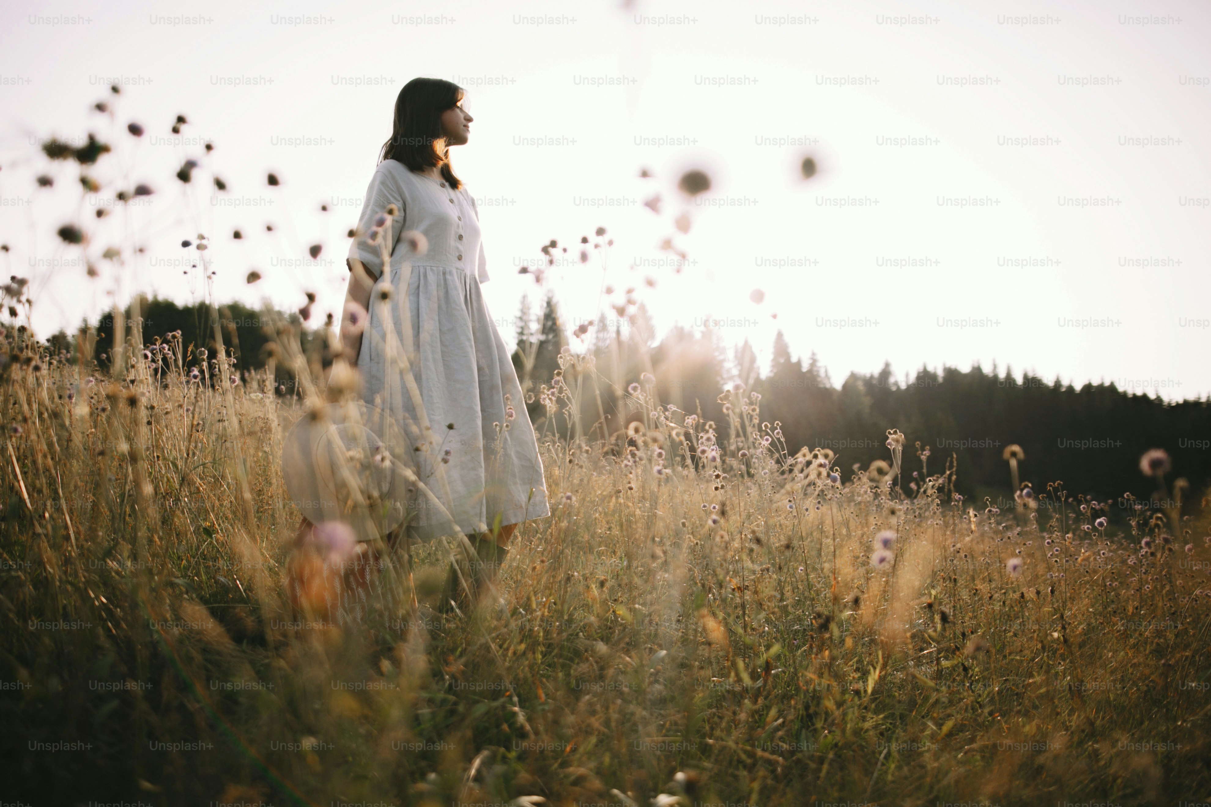 Stylish girl in rustic dress standing among wildflowers and herbs in sunny meadow  in mountains. Boho woman relaxing in countryside at sunset, simple life. Atmospheric image. Space text