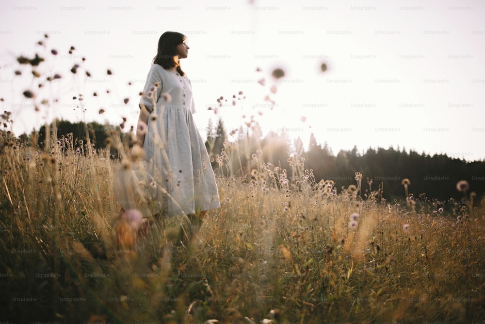 Menina elegante no vestido rústico em pé entre flores silvestres e ervas no prado ensolarado nas montanhas. Mulher Boho relaxando no campo ao pôr do sol, vida simples. Imagem atmosférica. Texto com espaço