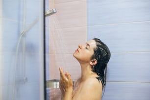 Giovane donna felice che fa la doccia a casa o nel bagno dell'hotel. Bella ragazza bruna che si lava i capelli e si gode il tempo di relax. Igiene del corpo e della pelle, concetto di stile di vita