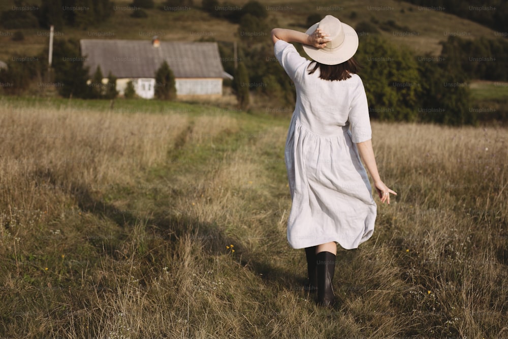 Muchacha elegante en vestido de lino y sombrero caminando en la hierba del campo soleado en el pueblo en las montañas. Mujer boho relajándose en el campo, vida rústica sencilla. Imagen atmosférica. Texto espaciado