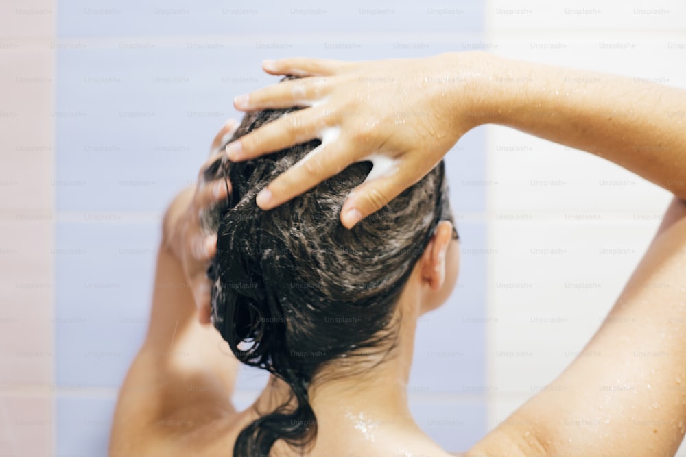 シャンプーで髪を洗う若い幸せな女性、泡の接写で手。シャワーを浴びてリラックスタイムを楽しんでいる美しいブルネットの女の子の背中。体と髪の衛生、ライフスタイルのコンセプト