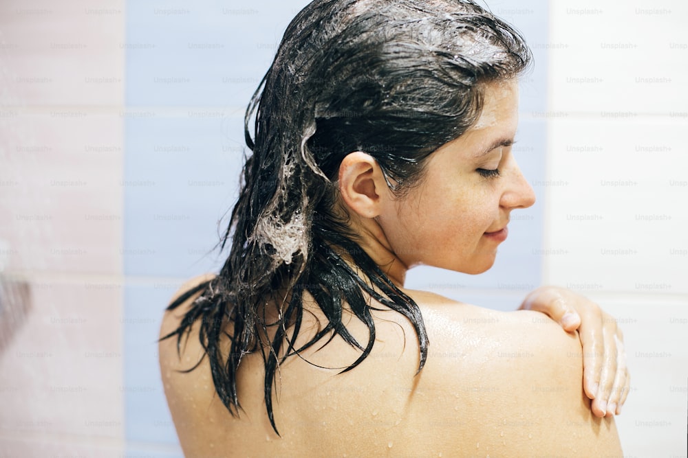Junge glückliche Frau, die ihre Haare mit Shampoo wäscht, nasses Haar mit Schaum. Rückseite des schönen brünetten Mädchens, das duscht und die Zeit genießt. Körper- und Haarhygiene, Lifestyle-Konzept. Text im Leerzeichen