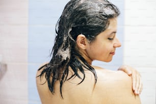 Young happy woman washing her hair with shampoo, wet hair with foam. Back of beautiful brunette girl taking shower and enjoying time. Body and hair hygiene, lifestyle concept. Space text