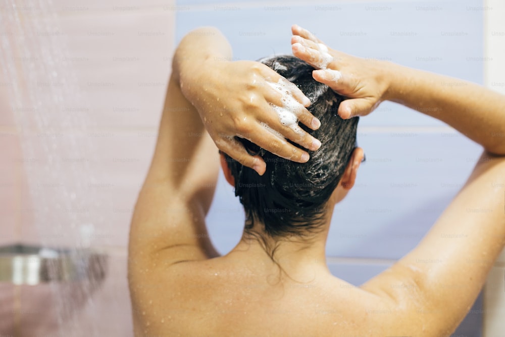 Young happy woman washing her hair with shampoo, hands with foam closeup. Back of beautiful brunette girl taking shower and enjoying relax time. Body and hair hygiene, lifestyle concept