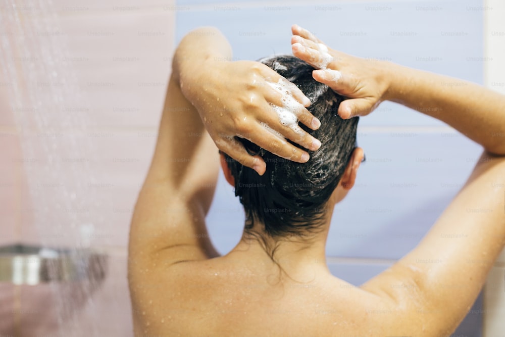 Junge glückliche Frau, die ihre Haare mit Shampoo wäscht, Hände mit Schaumstoffnahaufnahme. Rückseite des schönen brünetten Mädchens, das duscht und die Entspannungszeit genießt. Körper- und Haarhygiene, Lifestyle-Konzept