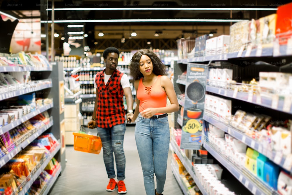 Handsome african american man with shopping basket holding hand of girlfriend while she looking for products in supermarket
