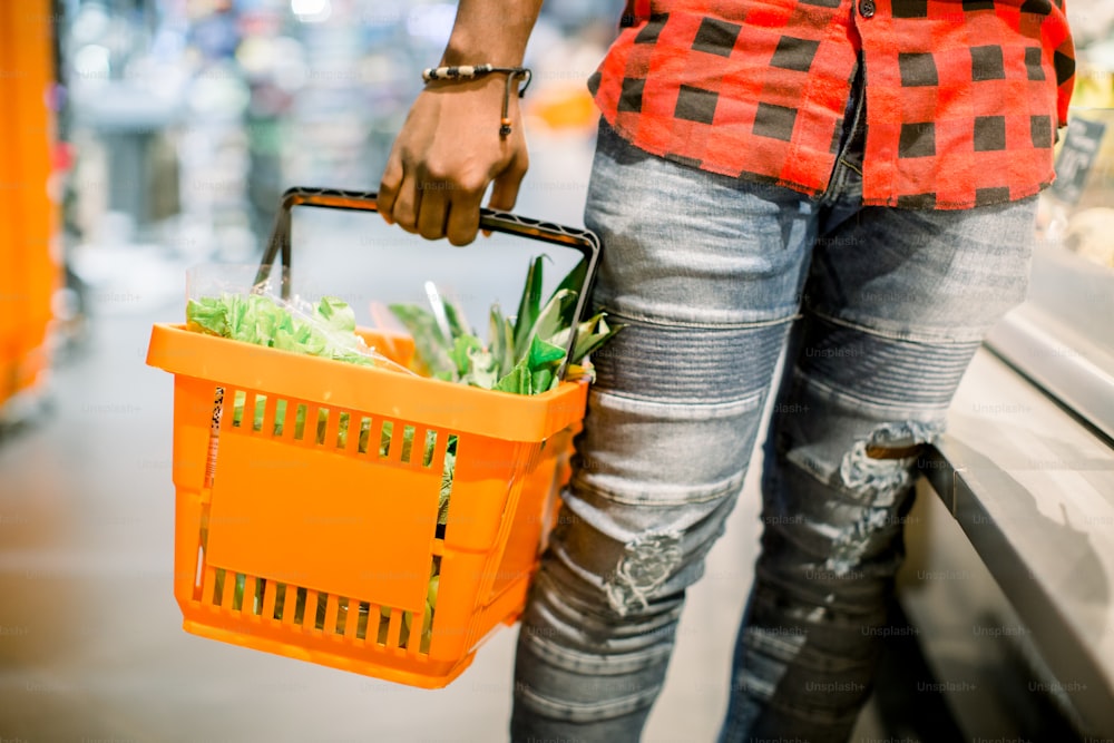 Joven africano comprando verduras y frutas en la sección de comestibles del supermercado. Un hombre negro elige verduras y frutas en el supermercado mientras sostiene una canasta de compras. Hombre comprando verduras en el supermercado.
