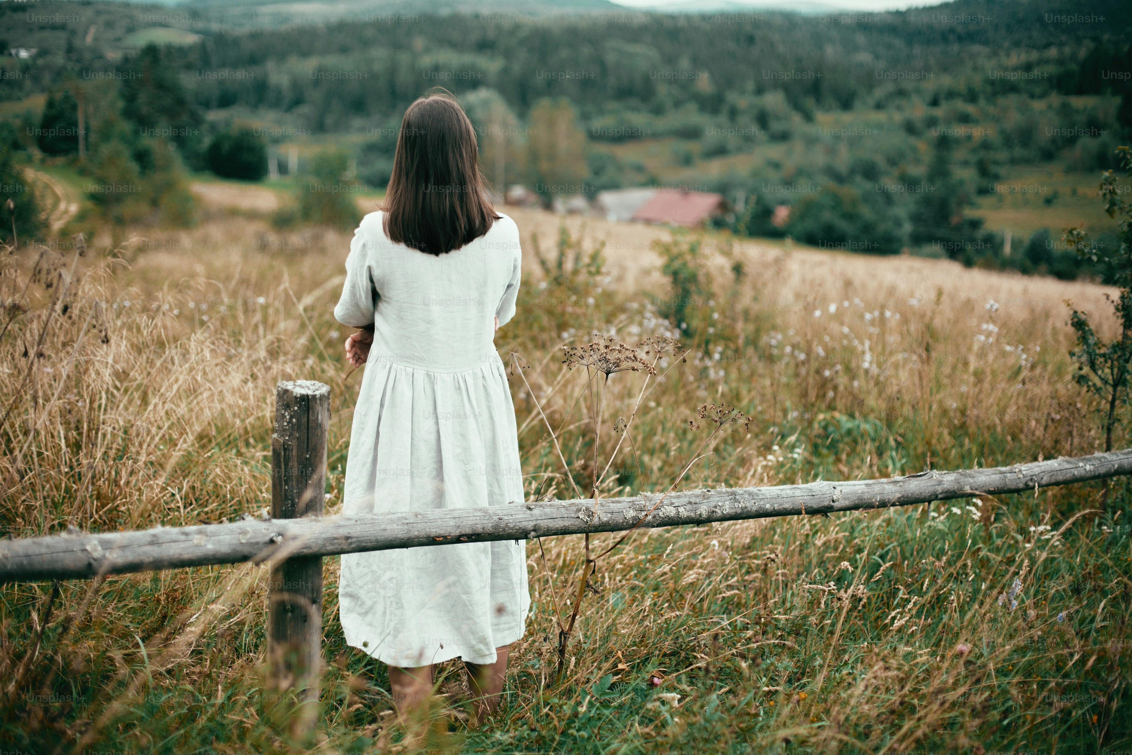 Stylish girl in linen dress standing at aged wooden fence among herbs and wildflowers, looking at field. Boho woman relaxing in countryside, simple slow life style.  Atmospheric image