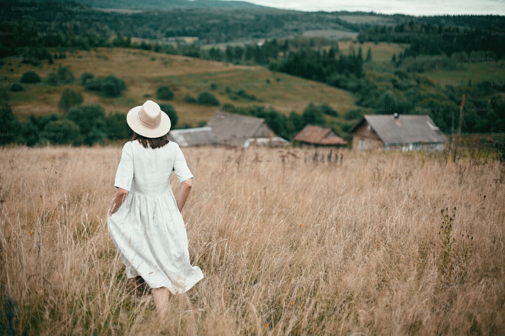 Stylish girl in linen dress and hat walking among herbs and wildflowers in field. Boho woman relaxing in countryside, simple slow life, amish style. Space for text. Atmospheric image