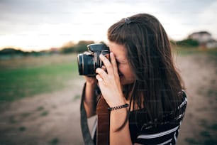 Stylish hipster girl taking photos on beach at sunset. Summer vacation. Portrait of  happy boho woman holding photo camera and making photos in evening time at  sea cliff