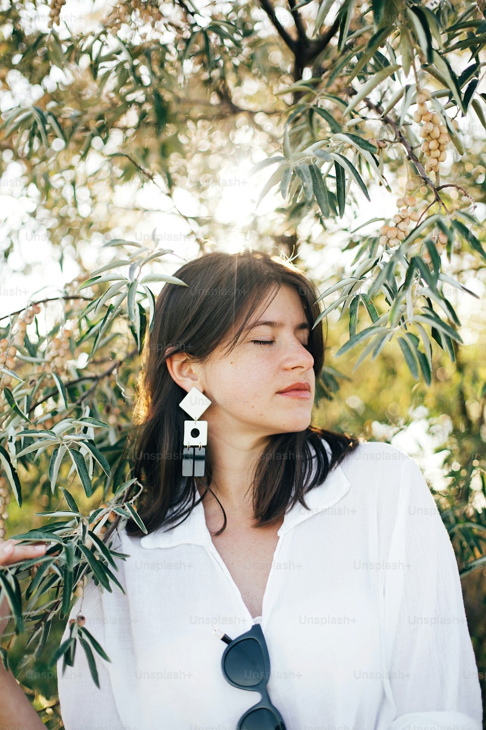 Portrait of young fashionable woman with modern earrings posing in  green olive branches in soft evening light, stylish boho girl relaxing on tropical island. Summer vacation.