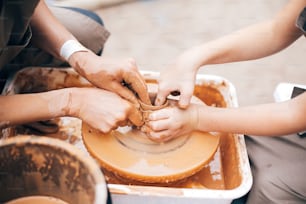 Hands of adult and child making pottery, working with wet clay closeup. Process of making bowl from clay on wheel with dirty hands. Handmade festival in summer park. Pottery workshop.