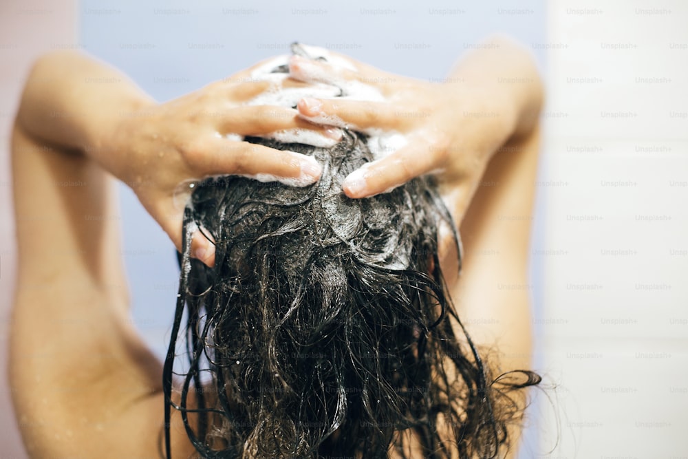 Junge glückliche Frau, die ihre Haare mit Shampoo wäscht, Hände mit Schaumstoffnahaufnahme. Rückseite des schönen brünetten Mädchens, das duscht und die Entspannungszeit genießt. Körper- und Haarhygiene, Lifestyle-Konzept