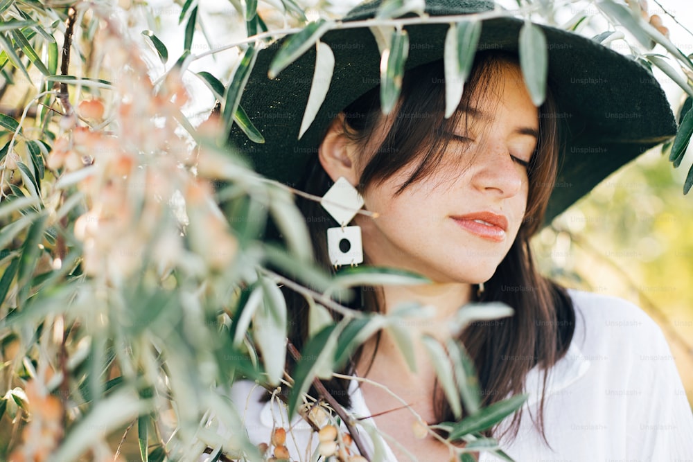 Stylish boho girl in hat and with modern earrings posing among green olive branches in soft evening light. Young fashionable woman relaxing on tropical island. Summer vacation. Space for text