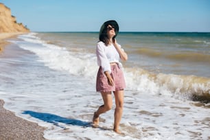 여름 방학. 열대 섬과 푸른 하늘에서 화창한 따뜻한 날에 바다 파도를 걷고 있는 행복한 젊은 보헤미안 여자. 텍스트를 위한 공간입니다. 해변에서 휴식을 취하고 웃고 있는 모자를 쓴 세련된 힙스터 소녀.
