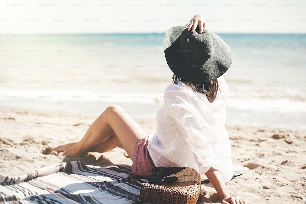 Chica hipster con estilo en sombrero sentada en la playa con bolsa de paja y bronceándose cerca de las olas del mar. Veraneo. Mujer boho feliz relajándose y disfrutando de un día cálido y soleado en el océano. Espacio para el texto