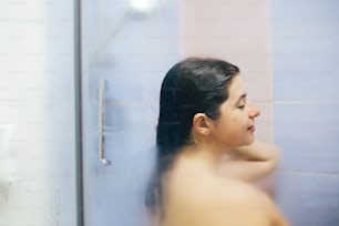 Young happy woman taking hot shower at home or hotel bathroom. Sensual portrait of beautiful brunette girl enjoying time in shower. Body and skin hygiene, lifestyle concept. Space text