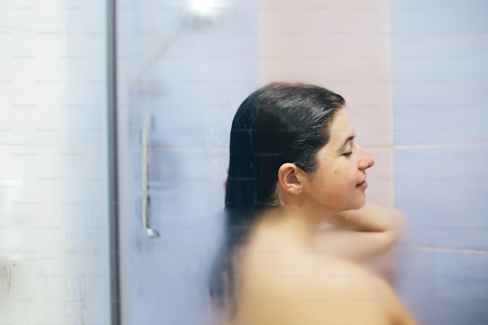 Young happy woman taking hot shower at home or hotel bathroom. Sensual portrait of beautiful brunette girl enjoying time in shower. Body and skin hygiene, lifestyle concept. Space text